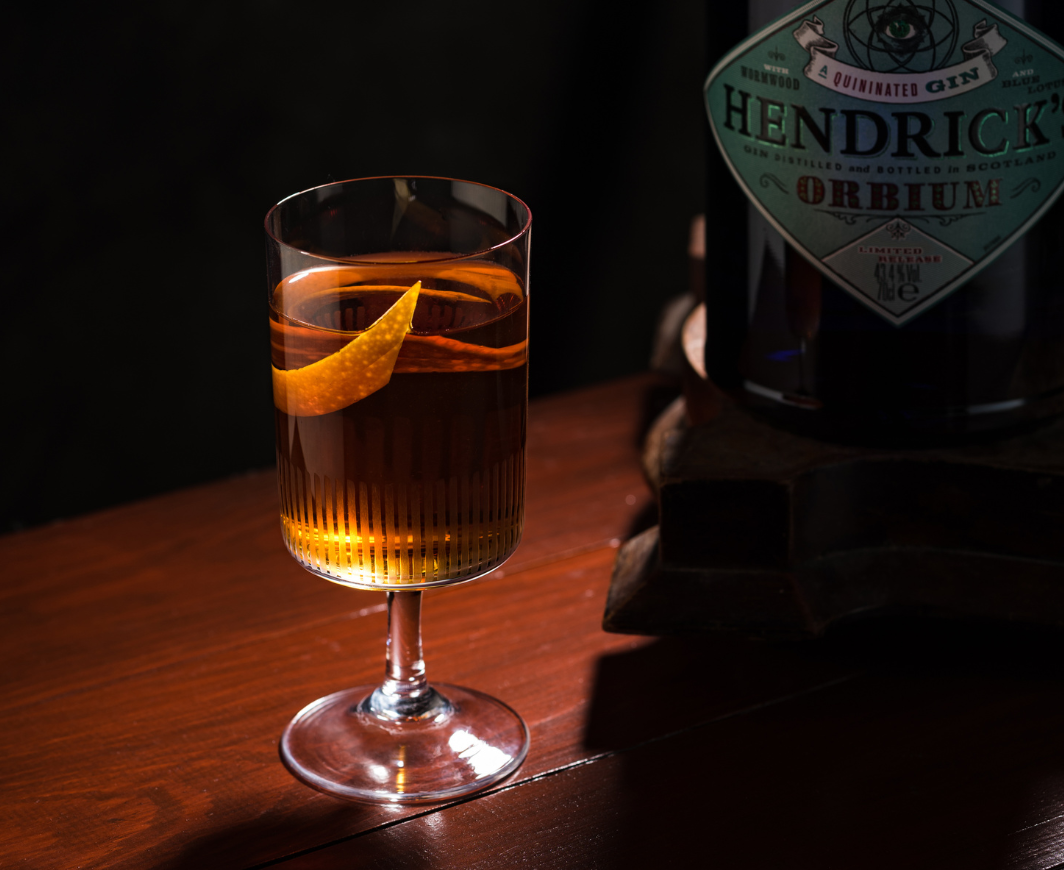 Eine Flasche Hendricks Orbium Martinez auf einem Podest auf einem Holztisch, davor ein Glas mit einem Orbium Martinez Cocktail