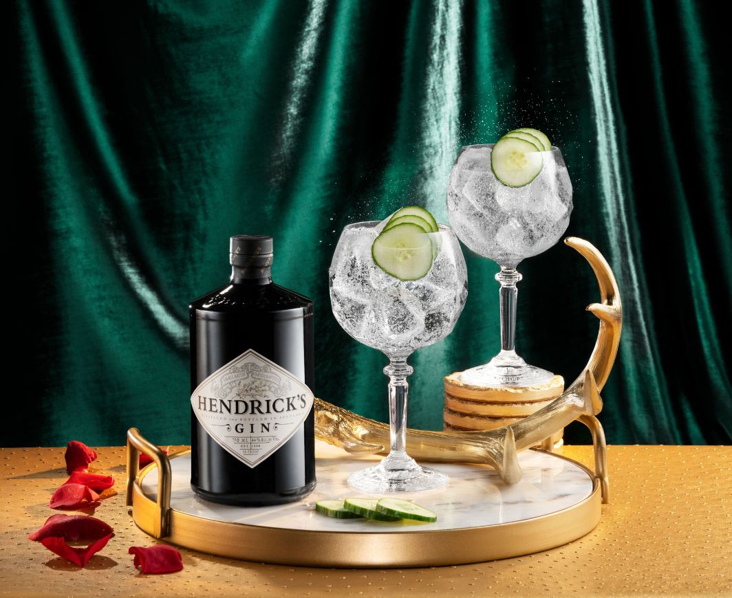 Eine Flasche Hendricks Gin auf mit zwei Gin Gläsern gefüllt mit Gin Tonic und garniert mit Gurkenscheiben auf eineme goldenen Tablett mit Marmorboden und einem goldenen Geweih als Dekoration, das ganze Ensemble auf einem goldenen Tisch mit grünem Samtvorhang im Hintergrund