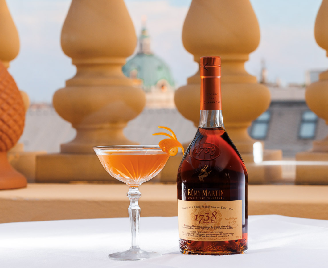 Eine Flasche Remy Martin 1738 nebem einem Glas mit einem Royal Sidecar Cocktail garniert mit einer Orangenzeste auf einem weißen Tisch vor einer Dächerkulisse einer Stadt