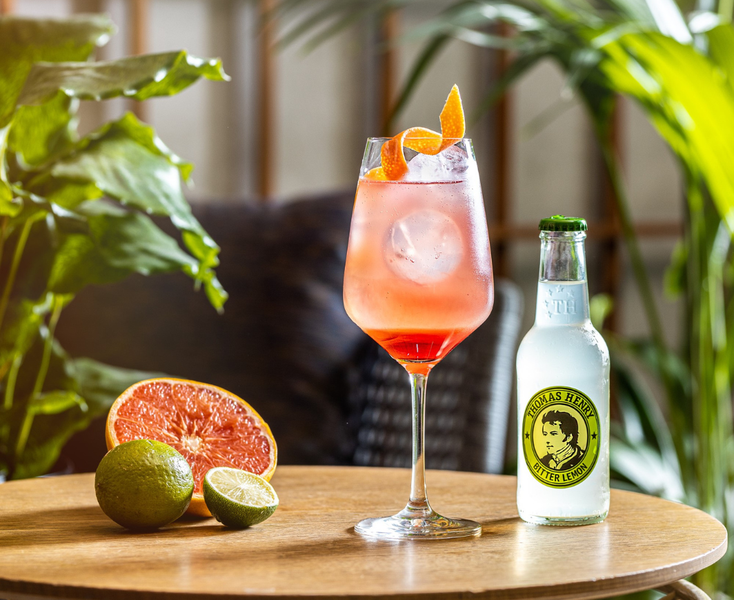 Ein Thomas Henry Amalfi Cocktail mit einer Flasche Thomas Henry Bitter Lemon und Zitronen und einer halben Grapefruit auf einem Holztisch vor einer Kulisse mit Pflanzen