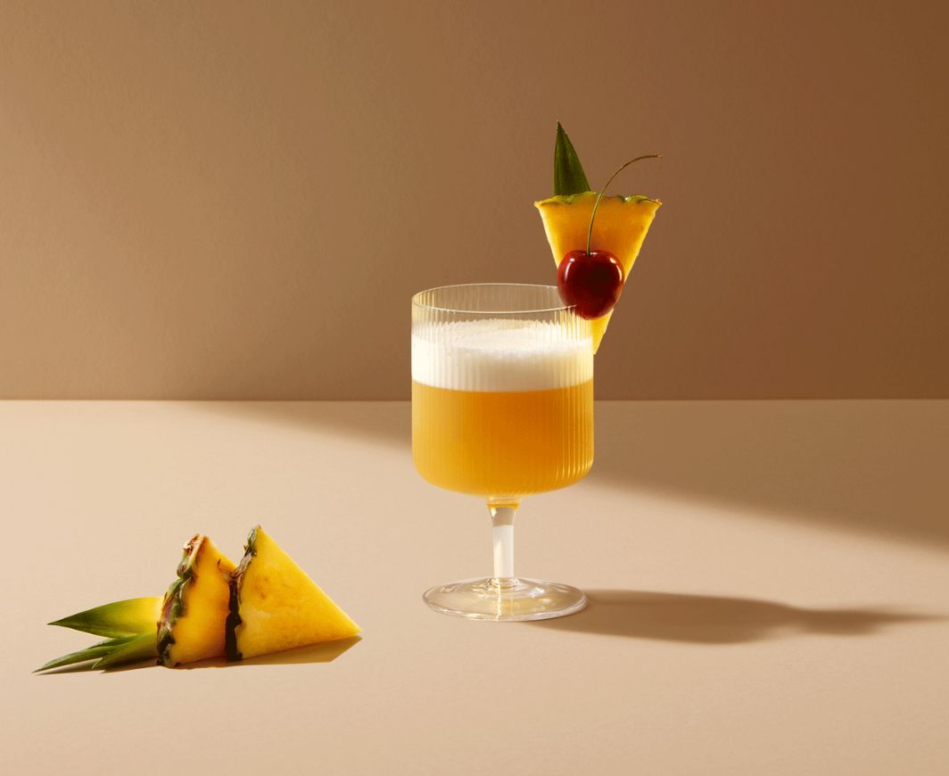 Ein Cocktail garniert mit Kirsche und Ananas auf neutralem Hintergrund und Boden, daneben zwei Scheiben Ananas