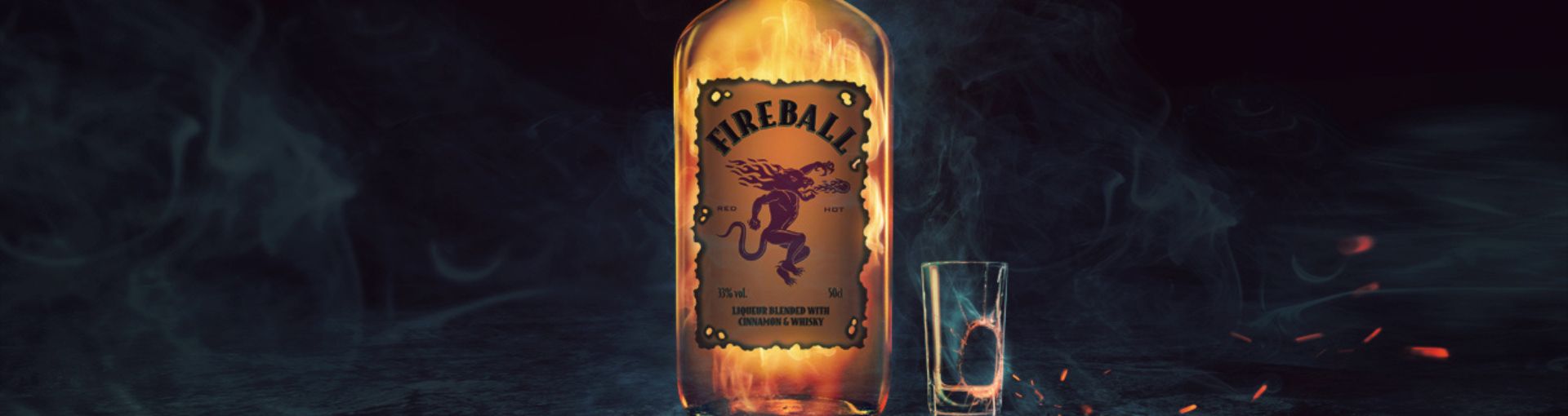 Fireball Whiskey Flasche in der Mitte des Bildes, daneben ein zerbrochenes Shotglas und leichter Rauch auf dem gesamten Bild