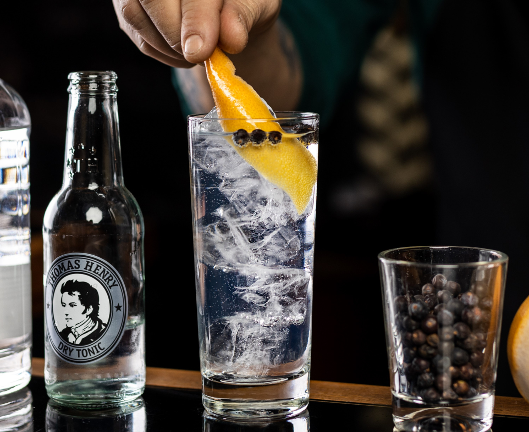 Ein Gin Tonic wird von einem Barkeeper mit einer Zitronenzeste garniert, rechts daneben ein Shotglas Wacholderbeeren und links daneben eine Flasche Thomas Henry Dry Tonic