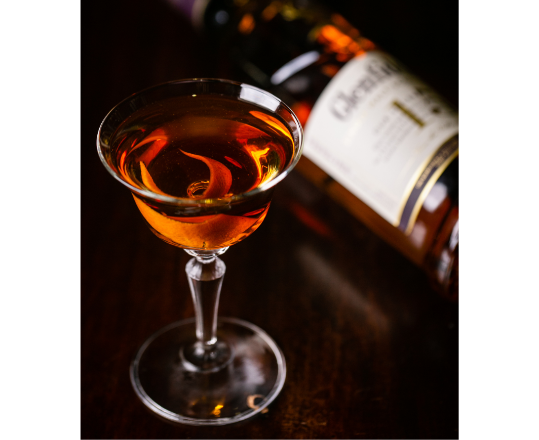 Ein Scotch Manhattan Cocktail in einem Glas auf einem Holztisch, dahinter eine verschwommene Flasche Glenfiddich 15 Jahre