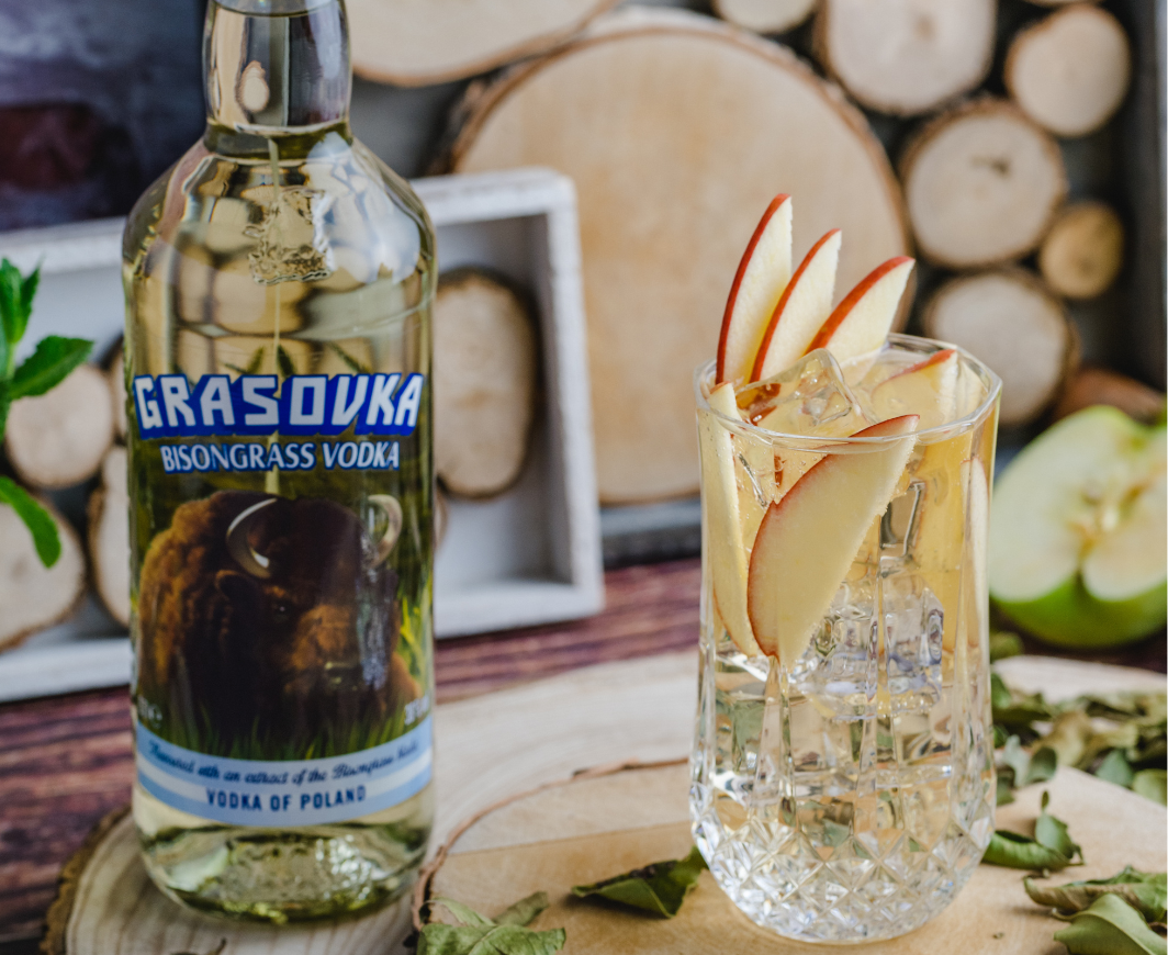 Eine Flasche Grasovka Bisongrass Vodka, daneben ein Cocktail garniert mit Eiswürfeln und Apfelscheiben auf einer Holzscheibe, alles vor einer Bank und einer Wand dekoriert mit Holzscheiben