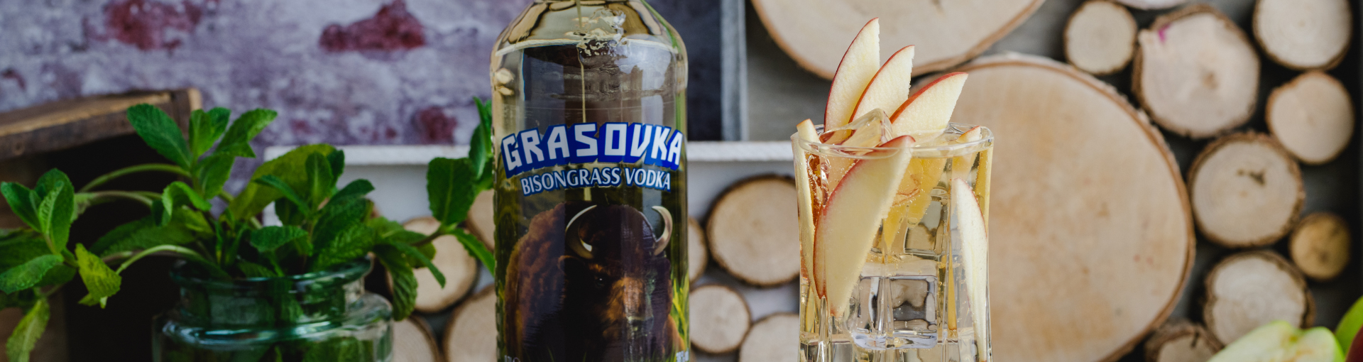 Eine Flasche Grasovka Vodka, daneben ein cocktailglas garniert mit Apfelscheiben, links daneben ein Glas mit Minze, im Hintergrund eine Wand dekoriert mit Holzscheiben