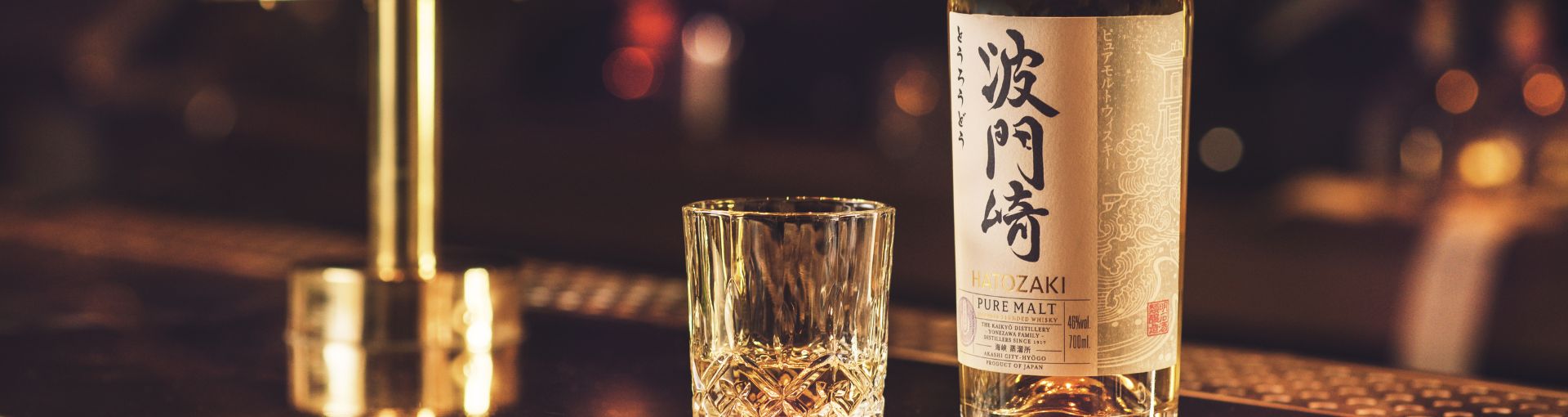 Ein Whiskey Glas gefüllt mit Hatozaki Whiskey und rechts daneben eine Flasche Hatozaki Whiskey, beides auf einer Bar mit einem goldenen Wasserhahn