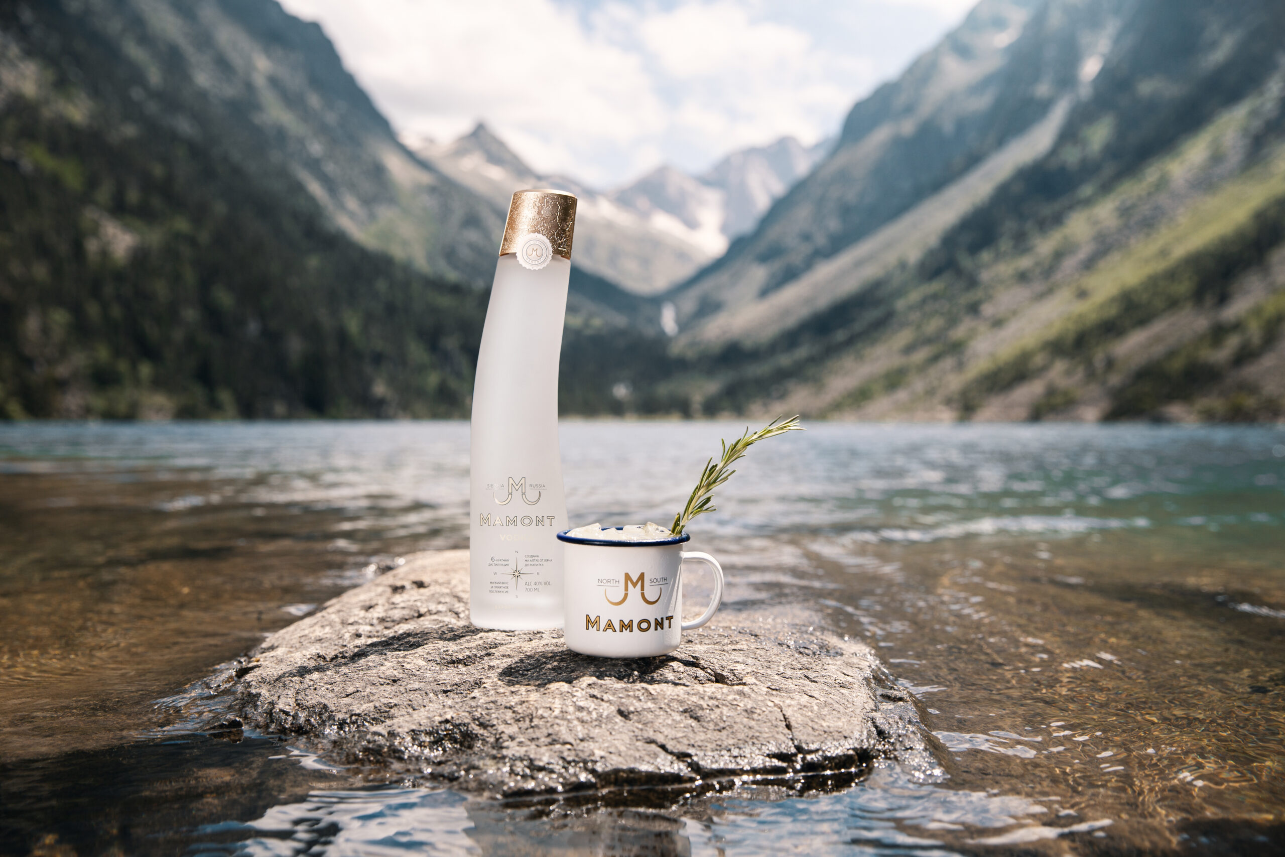 Mamont Flasche steht auf einem Stein in einem Fluss, daneben steht ein eingeschenktes Glas