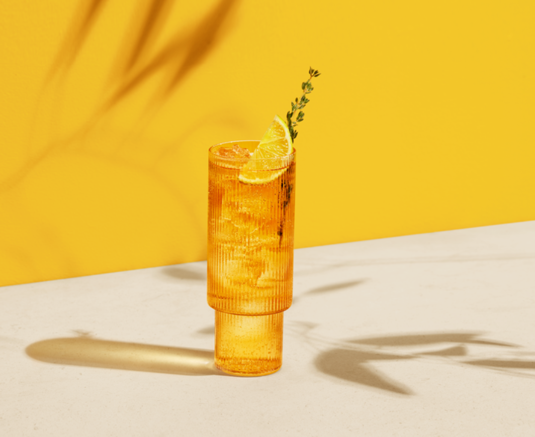 Longdrink Glas mit Mykonos Spritz und einer Zitronenscheibe und einem Rosmarin garniert auf einem weißen Tischg mit gelbem Hintergrund