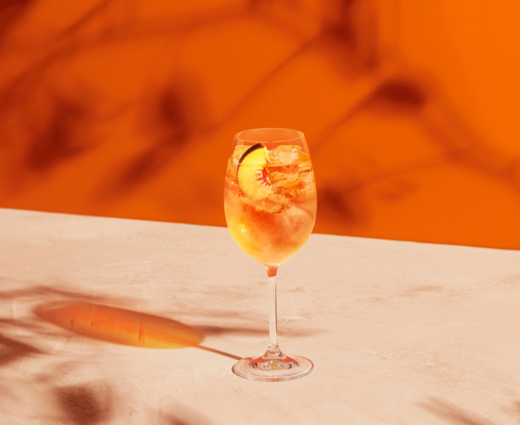 Weinglas mit einem Metaxa Pfirsich Spritz und einer Pfirsichscheibe garniert auf einem weißen Marmortisch mit orangenem Hintergrund