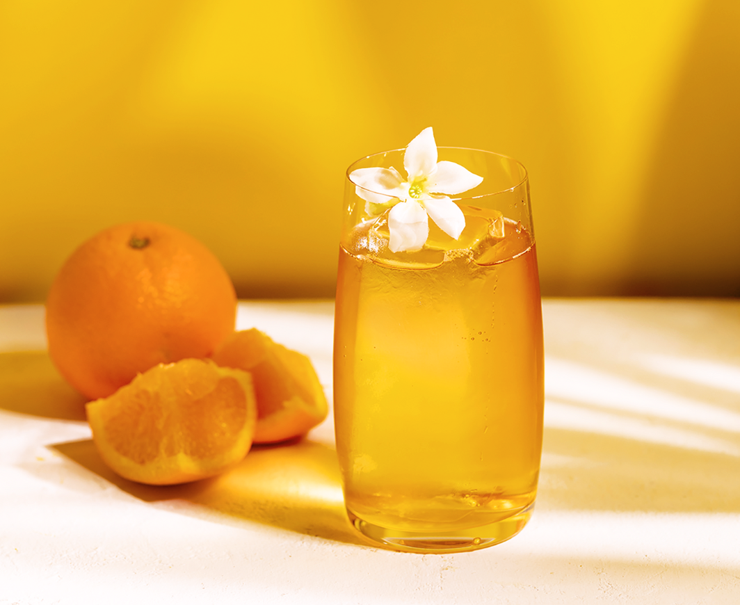 Ein Glas mit einem Metaxa Orange Tonic Cocktail garniert mit einer weißen Blume auf einem weißen Marmortisch, im Hintergrund eine Orange und Orangescheiben, alles vor einem hellorangen Hintergrund