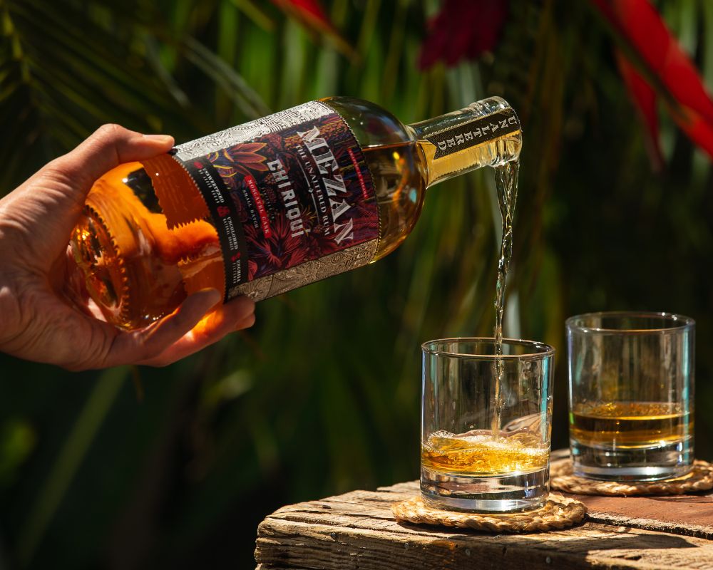 Eine Person gießt aus einer Flasche Mezan Whiskey den Whiskey ini ein Tumbler Glas, daneben steht noch ein Tumbler Glas mit Whiskey, beide Gläser stehen auf einer Holzkiste vor einem tropischen Hintergrund