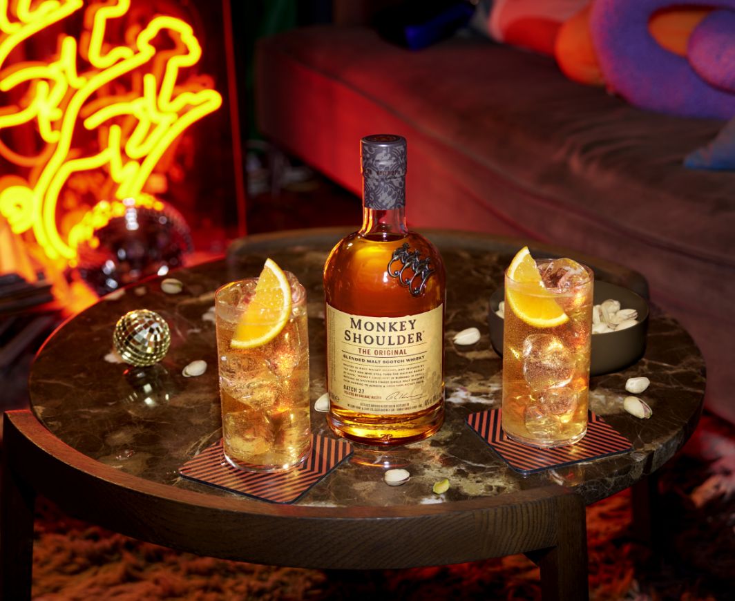 Eine Flasche Monkey Shoulder Whiskey auf einem Marmortisch mit zwei Longkdrinkgläsern gefüllt mit Cocktail davor und einer Schale Nüsse daneben so wie einer Discokugel, im Hintergrund ein Neonschild mit dem Motiv eines Affen