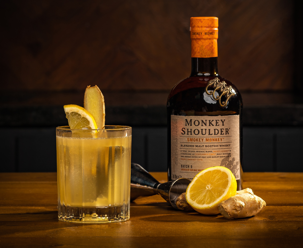 Eine Flasche Monkey Shoulder Smokey Monkey, davor ein Messbecher, Zitrone und Ingwer und links daneben ein Penicillin Serve Cocktail garniert mit einer Zitronenscheibe und einer Scheibe Ingwer, alles auf einem Holztisch vor einer Holzwand