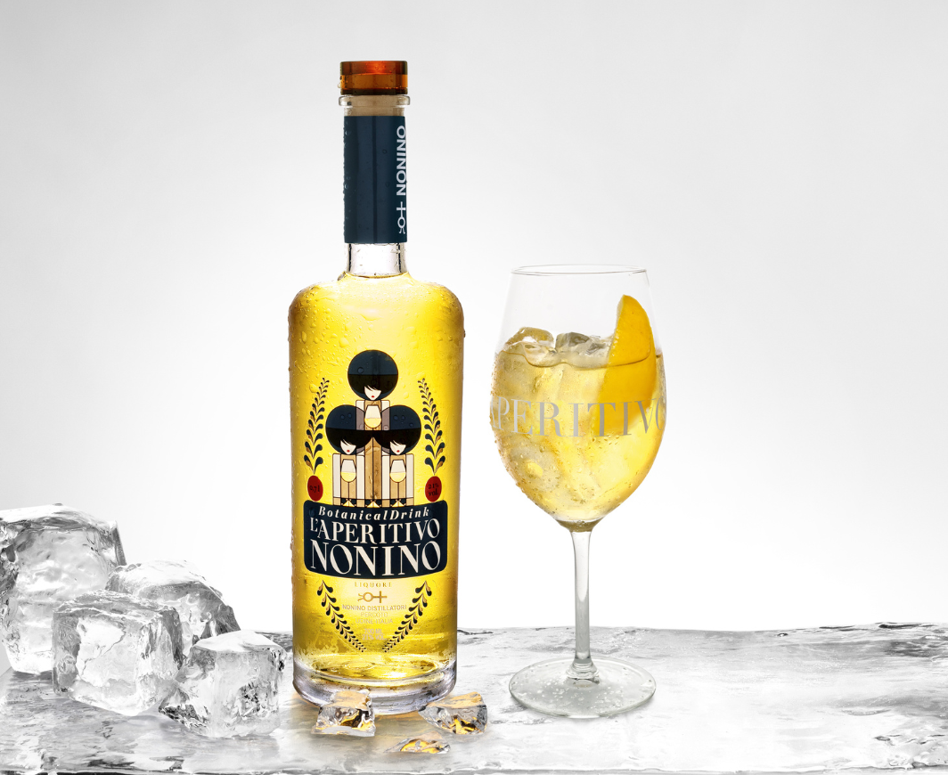 Eine Flasche L'Aperitivo Nonino eisgekühlt, rechts daneben ein Weinglas mit einem Cocktail und einer Zitronenteste garniert, links daneben Eiswürfel, alles angerichtet auf einem großen Eisblock vor neutralem Hintergrund