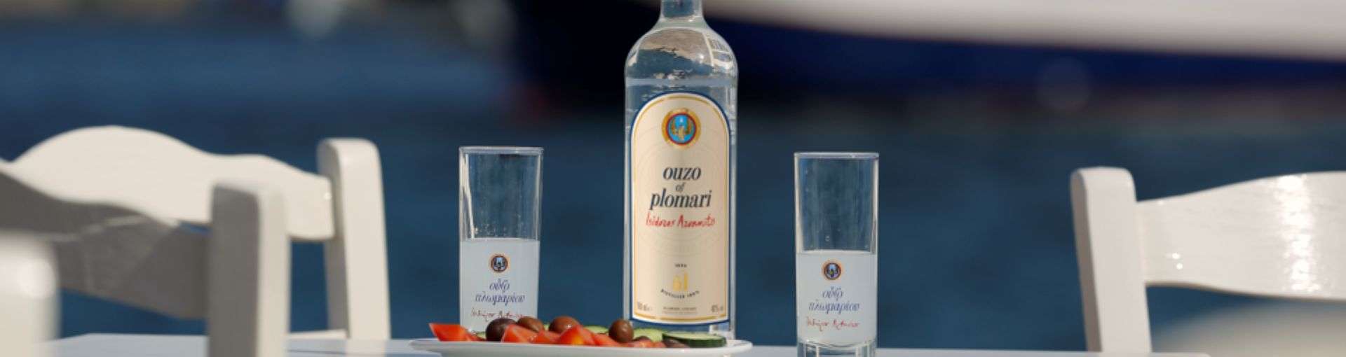 Ouzo of Plomari Flasche in der Mitte des Bildes, links und rechts daneben ein Glas für Ouzo und davor ein Teller mit Tomate, Oliven und Gurkenscheiben, alles steht auf einem Tisch mit weißen Sesseln darum und im Hintergrund das Meer mit einem blauen Boot