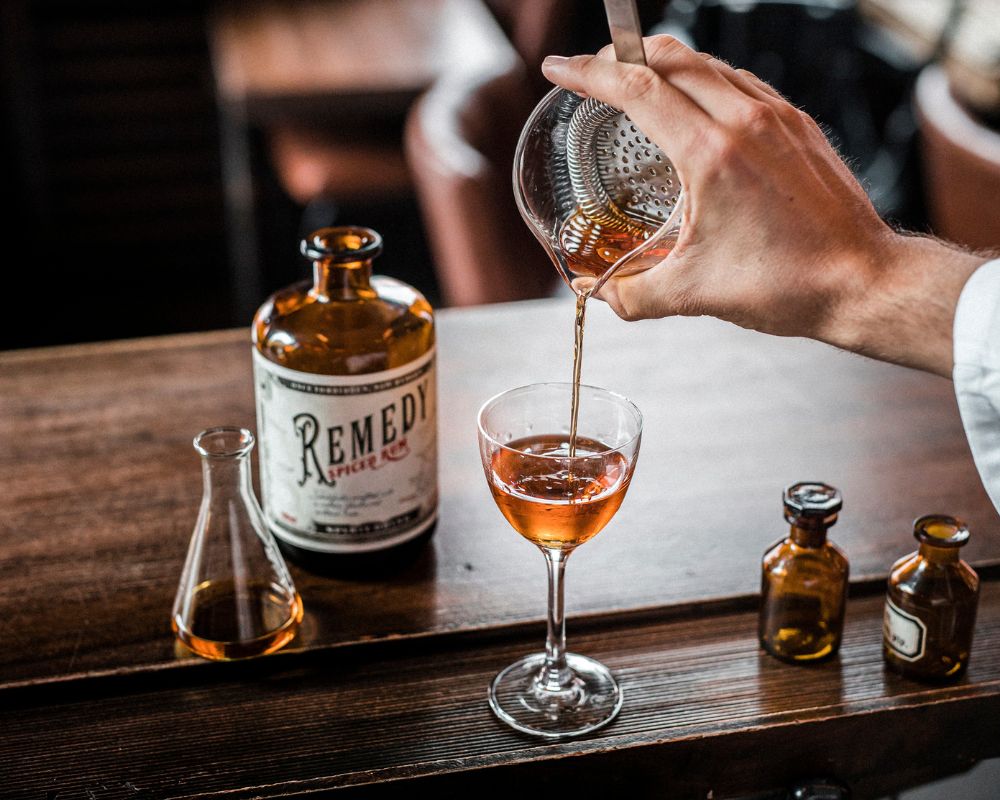 Eine Person die einen Cocktail in ein kleines Weinglas abseiht das auf einem Holztisch steht, dahinter eine Flasche Remedy Rum und daneben zwei kleine Flaschen