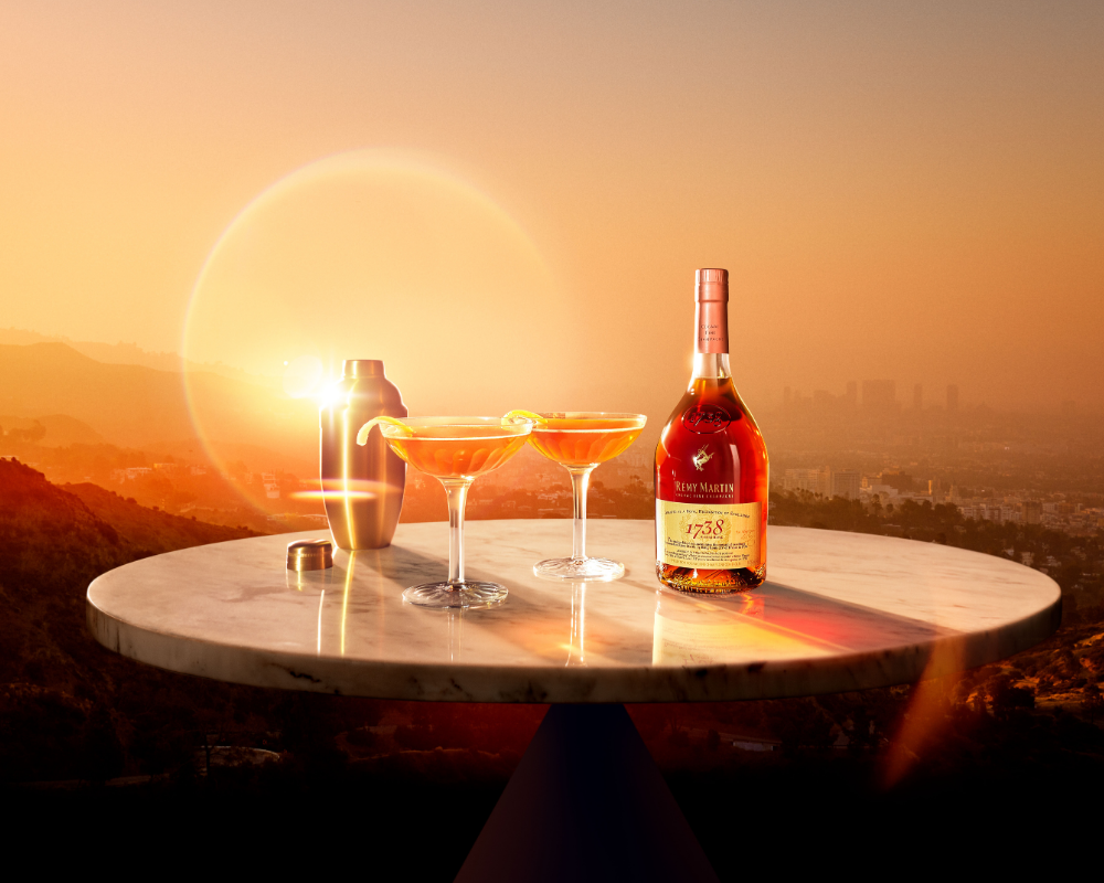 Eine Flasche Remy Martin 1738 mit 2 Gläsern gefüllt mit Cocktail und einem Cocktailshaker auf einem Marmortisch vor einer Skyline einer Stadt mit Sonnenaufgang