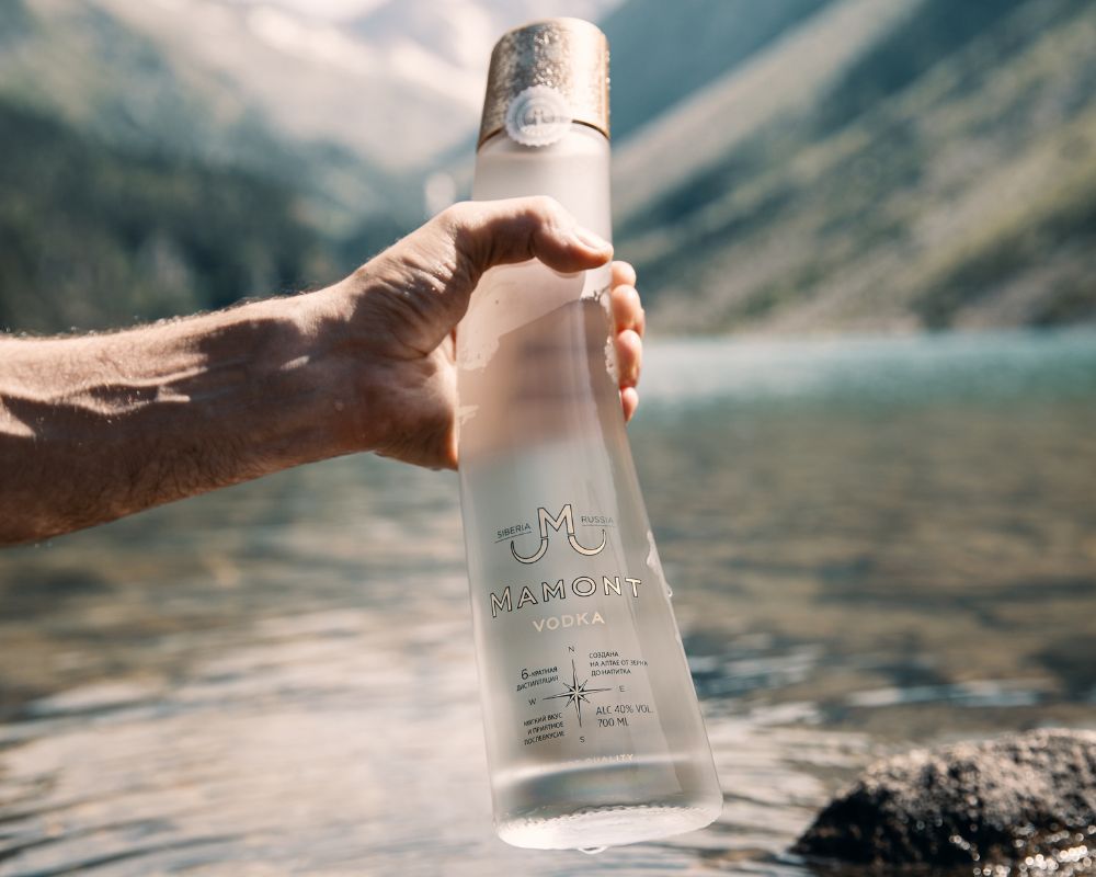 Person hält eine Flasche Mamont Vodka in der Hand, im Hintergrund sieht man einen Fluss und Berge