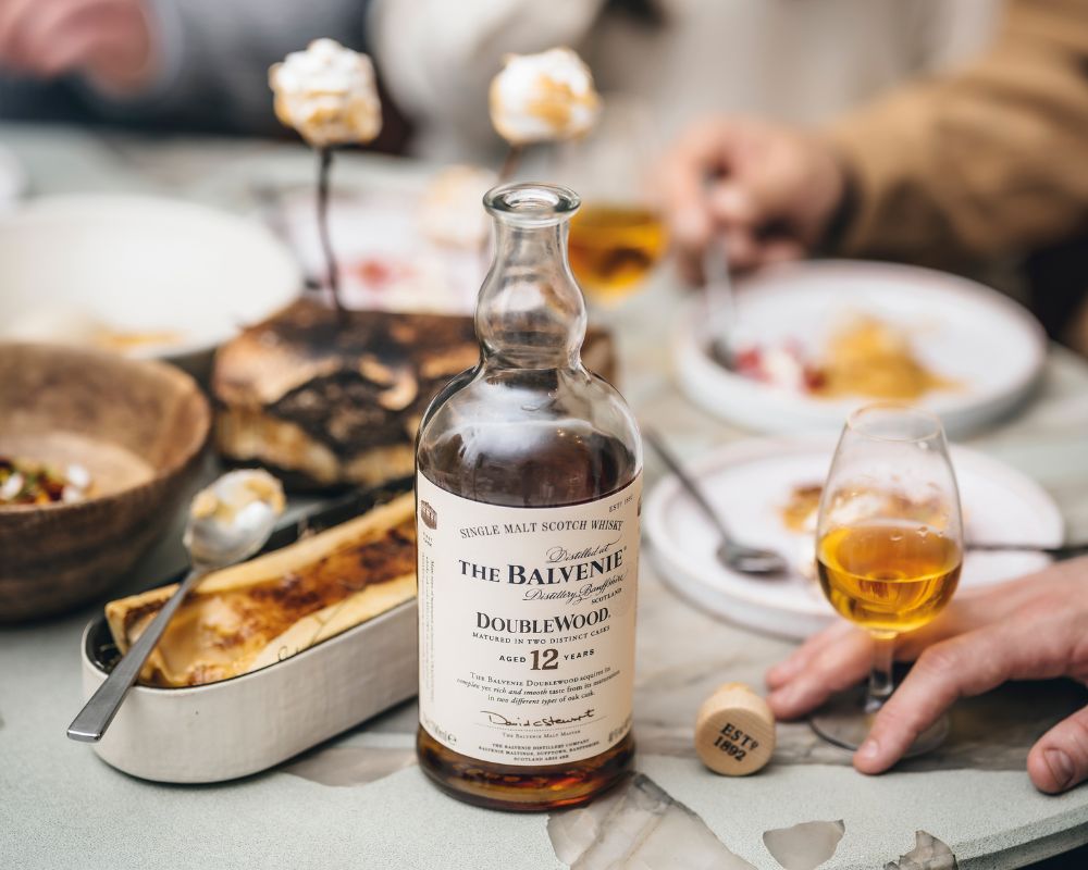 Balvenie steht auf gedecktem Tisch, eine Person hält ein Whiskey Glas fest, eine andere sieht man im Hintergrund