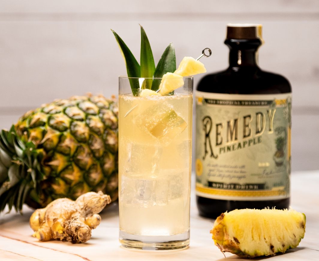 Eine Flasche Remedy Pinapple steht am Tisch neben einer Ananas und einem Drink