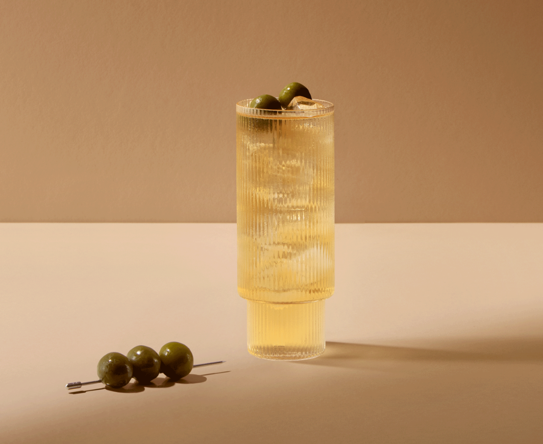 Ein Balvenie Speyside Tonic Cocktail in einem Longdrink Glas garniert mit Oliven, davor ein Spieß mit drei Oliven, alles auf einem neutralen Boden mit hellbraunem Hintergrund