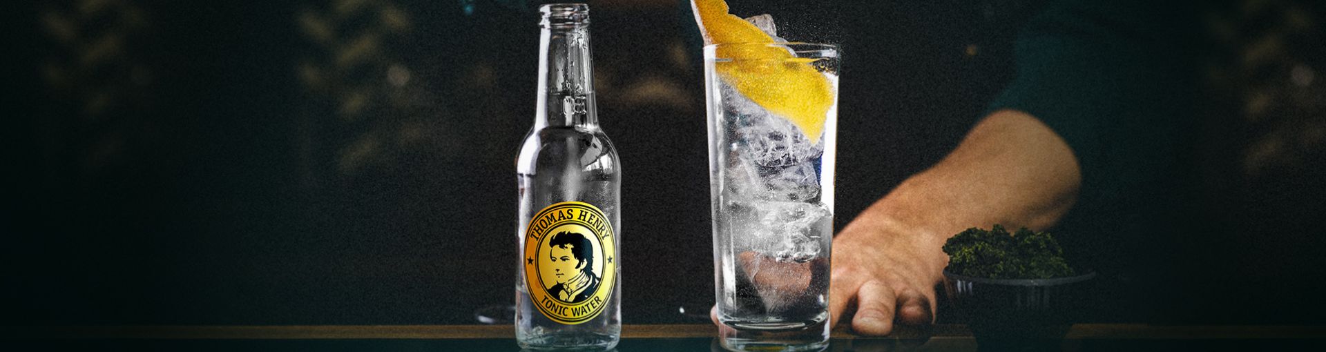 Cocktail im Longdrinkglas garniert mit Zitronenzeste, daneben eine Flasche Thomas Henry Tonic Water, rechts neben dem Longdrinkglas eine Schüssel Petersilie und im HIntergrund eine Person die den Cocktail auf den Tisch schiebt