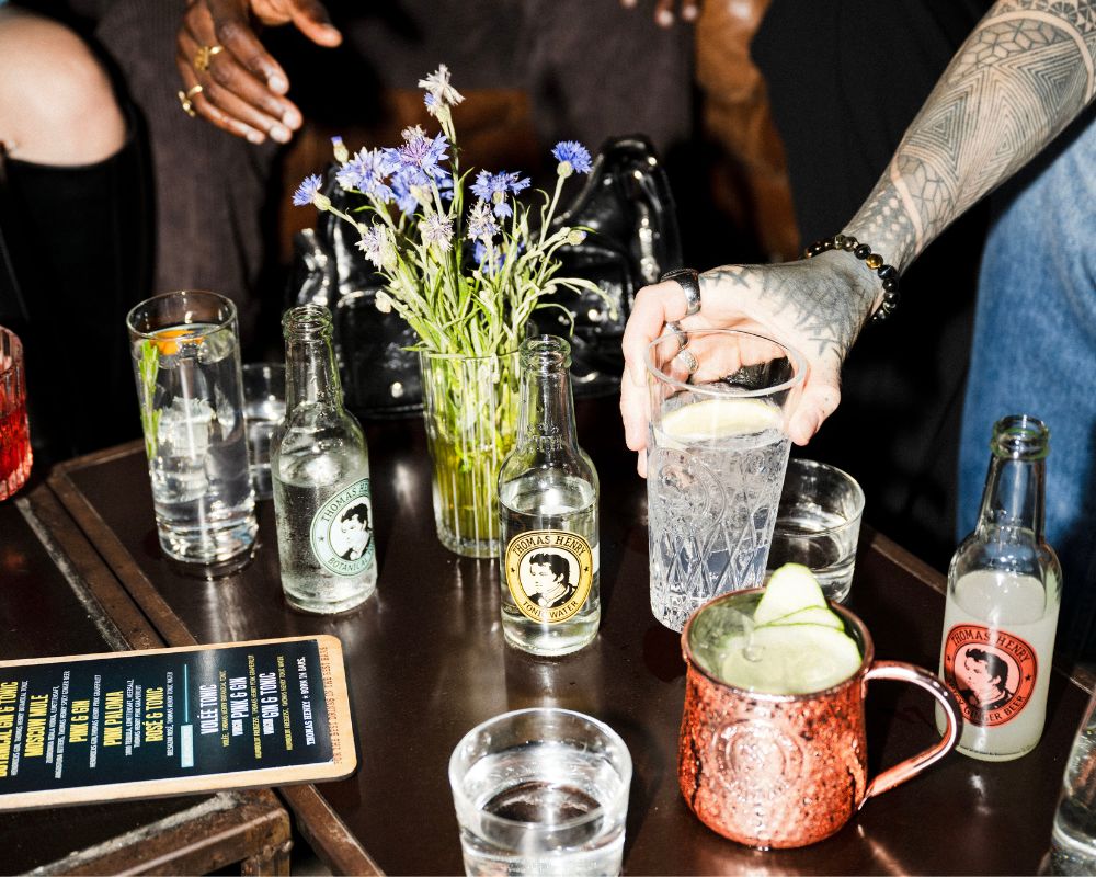 Cocktails gemixxt mit Thomas Henry Flaschen auf einem Holztisch mit Blumen und Barkarte als Dekoration, um den Tisch herum eine Personengruppe und eine Person mit Tattoos am Arm greift einen Cocktail