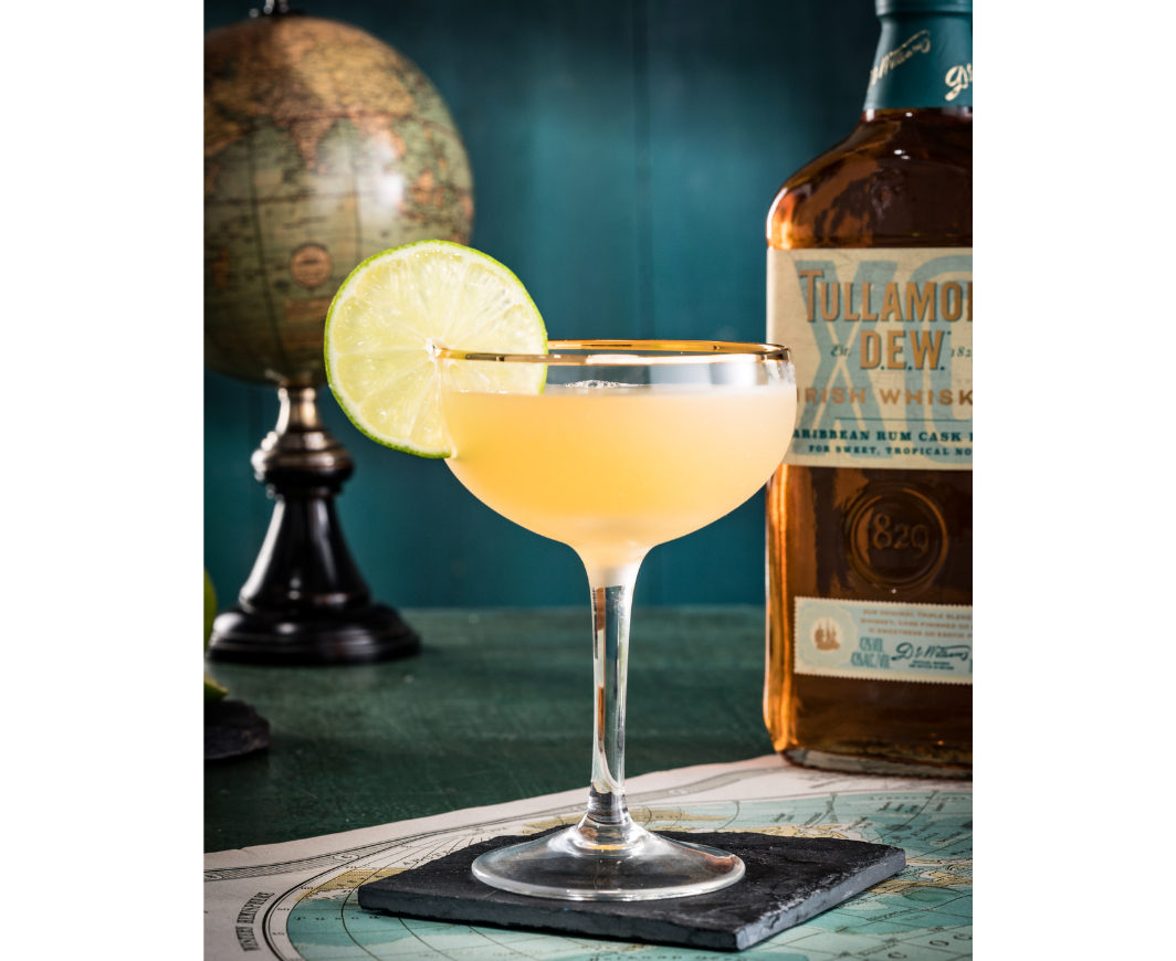 Eine Cocktailschale gefüllt mit einem Tullamore Daiquiri Cocktail garniert mit einer Limettenscheibe auf einer Schiefersteinplatte auf einer Weltkarte, dahinter eine Flasche Tullamore D.E.W. und weiter dahinter ein Glous